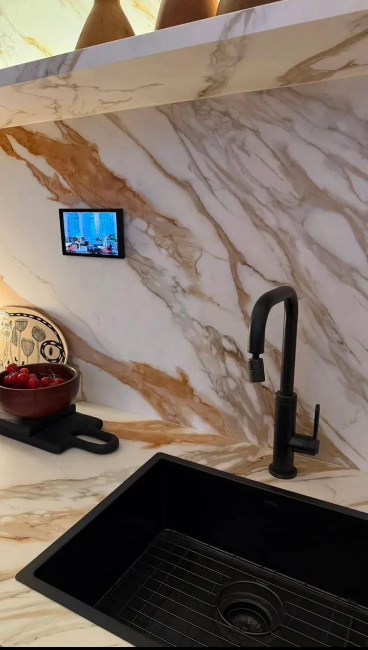Cozinha com bancada e parede de mármore branco com veios dourados, cuba e torneira pretas e um pequeno tablet embutido na parede.