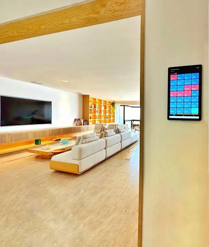 Sala ampla com sofá claro e estante iluminada, vista do corredor, com um tablet na parede mostrando as cenas da casa.